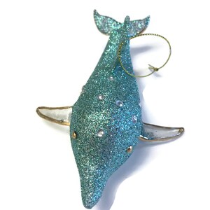 K&ouml;nnte beinhalten: Ein tealblauer glitzernder Delfin-Anh&auml;nger mit einer goldenen Schnur zum Aufh&auml;ngen. Der Delfin hat wei&szlig;e Glitzerakzente und goldene Details.