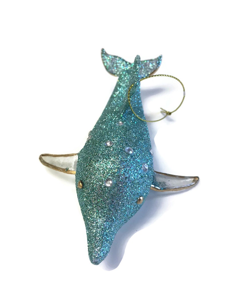 K&ouml;nnte beinhalten: Ein tealblauer glitzernder Delfin-Anh&auml;nger mit einer goldenen Schnur zum Aufh&auml;ngen. Der Delfin hat wei&szlig;e Glitzerakzente und goldene Details.