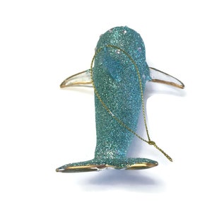 K&ouml;nnte beinhalten: Ein tealblauer Glas-Anh&auml;nger in Form eines Delfins mit goldenen Glitzer und wei&szlig;en Perlenakzenten. Der Anh&auml;nger hat eine goldene Schnur zum Aufh&auml;ngen.