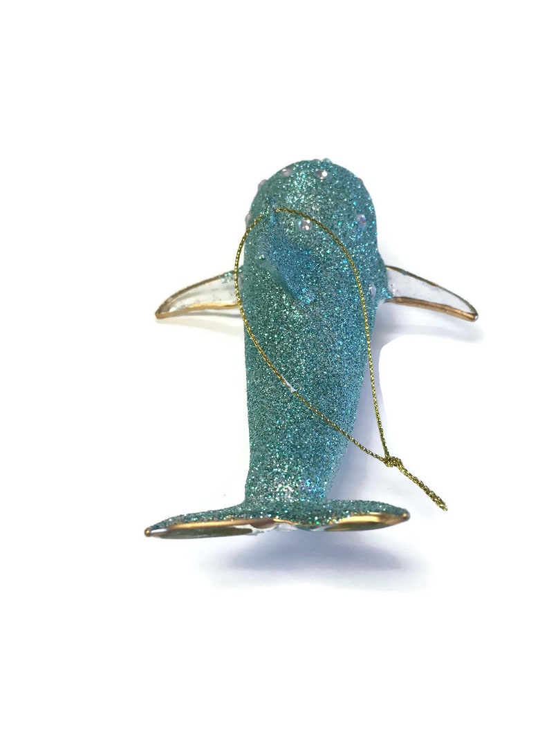 K&ouml;nnte beinhalten: Ein tealblauer Glas-Anh&auml;nger in Form eines Delfins mit goldenen Glitzer und wei&szlig;en Perlenakzenten. Der Anh&auml;nger hat eine goldene Schnur zum Aufh&auml;ngen.