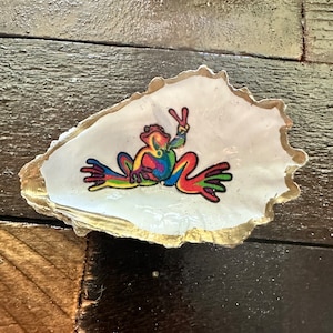 May include: A white oyster shell with a gold rim, featuring a colorful frog with a peace sign. The frog is painted in shades of red, orange, yellow, green, blue, and purple.