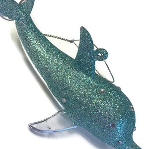 K&ouml;nnte beinhalten: Ein tealblauer Glitzer-Delfin-Anh&auml;nger mit silbernen Akzenten. Der Delfin hat eine durchsichtige Plastikflosse und eine silberne Schnur zum Aufh&auml;ngen.