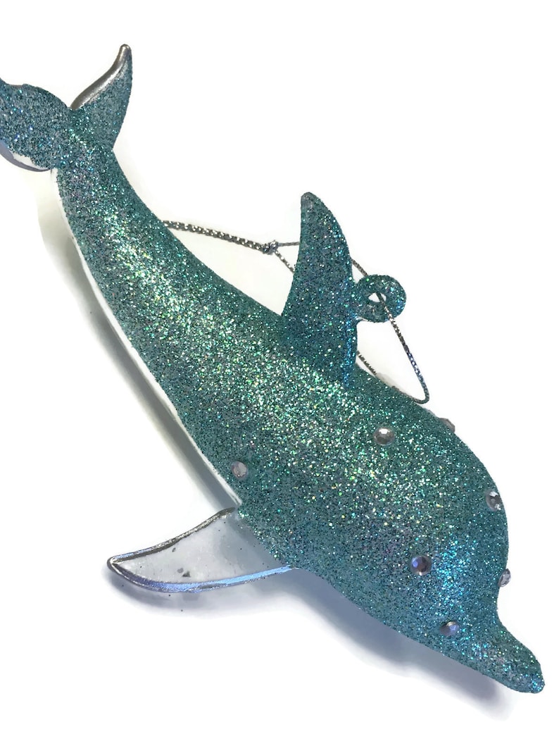 K&ouml;nnte beinhalten: Ein tealblauer Glitzer-Delfin-Anh&auml;nger mit silbernen Akzenten. Der Delfin hat eine durchsichtige Plastikflosse und eine silberne Schnur zum Aufh&auml;ngen.