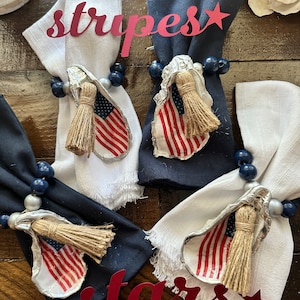 May include: Four napkin rings made from oyster shells with a small American flag and a jute tassel. The napkin rings are on white and navy blue napkins. The words "stripes" and "stars" are written in red.