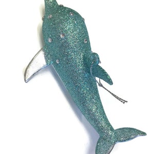 K&ouml;nnte beinhalten: Ein tealblauer glitzernder Delfin-Weihnachtsbaumschmuck mit silbernen Akzenten. Der Delfin hat eine silberne Schwanzflosse und eine silberne R&uuml;ckenflosse. Der Anh&auml;nger hat eine silberne Schnur zum Aufh&auml;ngen.