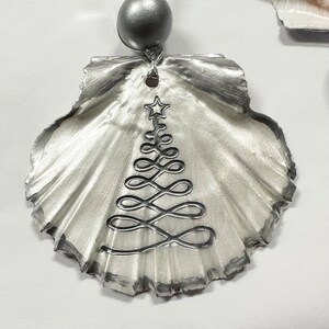 May include: A silver seashell ornament with a white Christmas tree design. The ornament has a silver bead at the top for hanging.