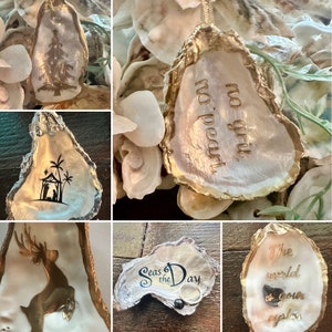 May include: A collection of oyster shells with various designs and sayings. The shells are painted gold and white and feature designs such as a Christmas tree, a nativity scene, a deer, and the phrase "Seas the Day". One shell has the saying "No grit, no pearl".