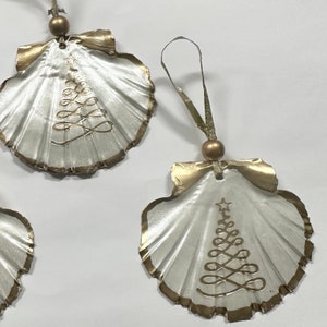 May include: Three white and gold Christmas ornaments shaped like seashells with a gold Christmas tree design.