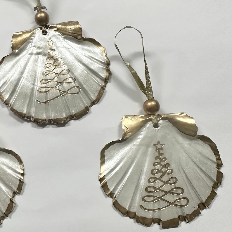May include: Three white and gold Christmas ornaments shaped like seashells with a gold Christmas tree design.