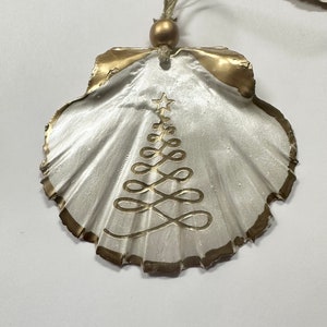 May include: A white seashell ornament with a gold trim and a gold painted Christmas tree design. The ornament has a gold bead and a green ribbon for hanging.
