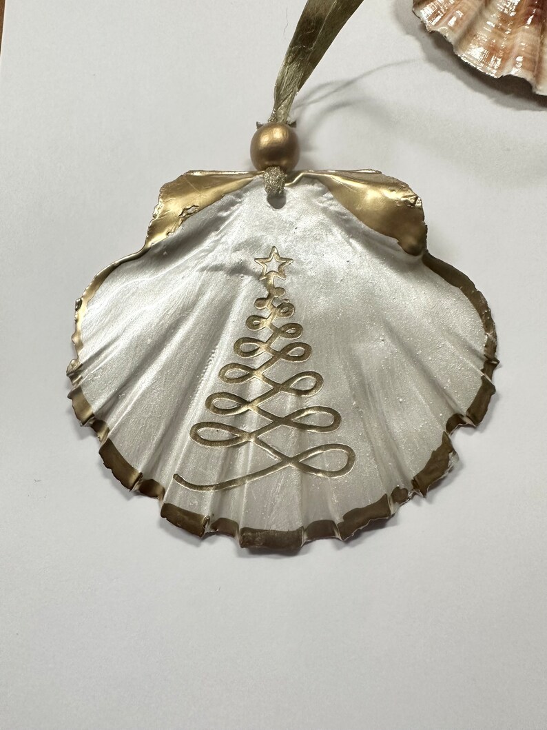 May include: A white seashell ornament with a gold trim and a gold painted Christmas tree design. The ornament has a gold bead and a green ribbon for hanging.