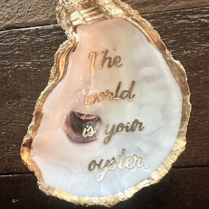 May include: A white oyster shell with a gold rim, the inside is painted with the words "The world is your oyster."