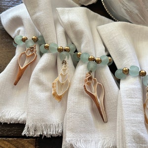 May include: Four white linen napkins with fringed edges are tied with rings of light blue and gold glass beads. Each ring has a seashell charm attached.