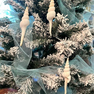 May include: Three white seashell ornaments hanging on a flocked Christmas tree with a blue tulle ribbon.