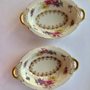 May include: Two oval-shaped serving bowls with gold trim and floral designs. The bowls have two handles on each side.
