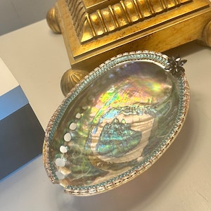 May include: A large iridescent abalone shell with a beaded trim and a small black accent. The shell is sitting on a white surface.