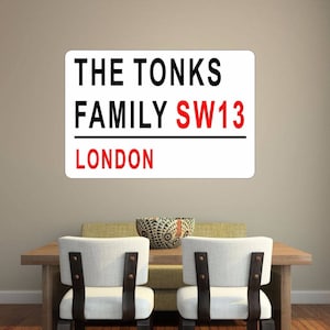 May include: White sign with black text that reads "THE TONKS FAMILY SW13 LONDON". The sign is mounted on a light beige wall above a wooden table with two white chairs.