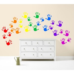 May include: A white dresser with seven drawers is positioned below a series of colorful handprints in a rainbow arc. The handprints are red, orange, yellow, green, blue, and purple. A green pitcher sits on the floor.