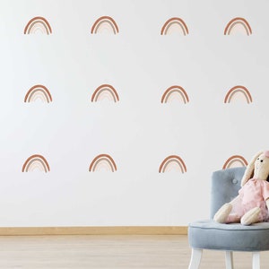 May include: A white wall adorned with brown and beige rainbow decals. A grey chair holds a stuffed bunny in a pink dress. Decorative pom-poms are on a stand in the background. The room has a wooden floor.