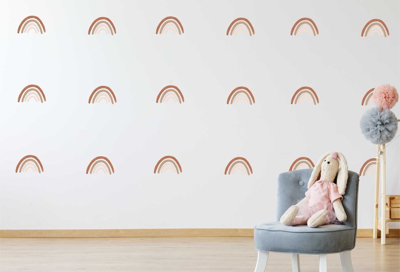 May include: A white wall adorned with brown and beige rainbow decals. A grey chair holds a stuffed bunny in a pink dress. Decorative pom-poms are on a stand in the background. The room has a wooden floor.