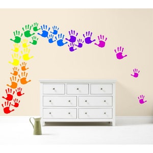 May include: A white dresser with multiple drawers is positioned below a wall decorated with colorful handprints. The handprints are arranged in a rainbow arc, with additional handprints scattered across the wall. A green pitcher sits on the floor.