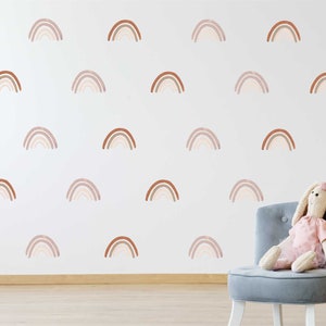 May include: A white wall decorated with multiple watercolour-style rainbow decals in shades of brown and beige. A plush bunny toy sits on a grey chair. Decorative pom-poms add a touch of whimsy to the room.