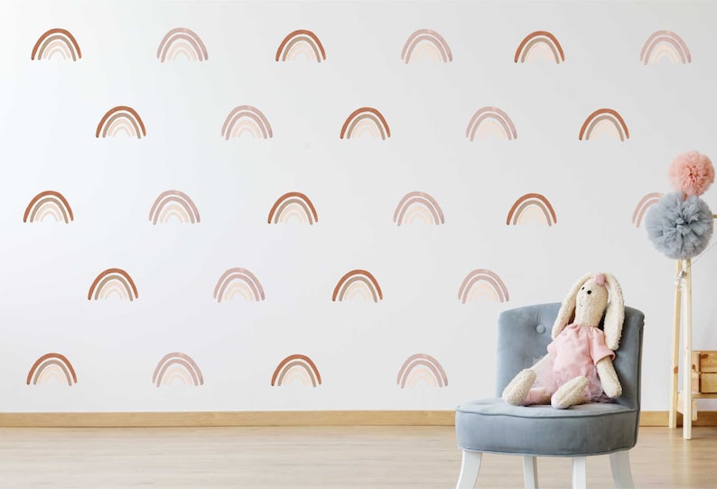 May include: A white wall decorated with multiple watercolour-style rainbow decals in shades of brown and beige. A plush bunny toy sits on a grey chair. Decorative pom-poms add a touch of whimsy to the room.