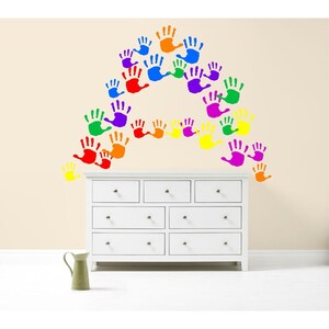 May include: A white dresser with multiple drawers is positioned below a wall decorated with colorful handprints. The handprints are arranged in an arch shape, using a variety of colors including red, orange, yellow, green, blue, and purple.