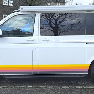 Könnte beinhalten: Ein weißer Transporter mit einem horizontalen Streifendekor an den unteren Seitenwänden in den Farben Gelb, Orange, Rosa und Grau. Das Fahrzeug verfügt über getönte Heckscheiben, silberne Leichtmetallfelgen und eine weiße ausziehbare Dachmarkise.