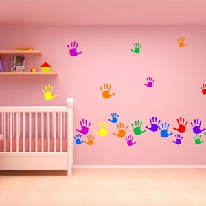 May include: A pink nursery with a white crib, a dresser, and shelves. Colorful handprints in yellow, orange, green, blue, purple, and red are on the pink wall. A teddy bear sits on the dresser.