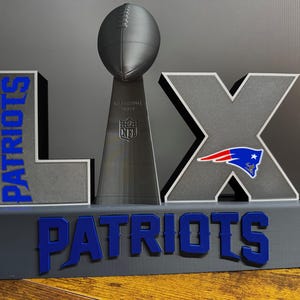 May include: A gray and blue decorative piece featuring the word "PATRIOTS" in blue, along with the Roman numeral "LIX" and a football-shaped trophy. The Patriots logo is visible on the "X".
