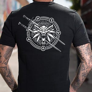 May include: A black short-sleeved t-shirt featuring a white graphic on the back. The design displays a stylized wolf head centered within a circular emblem, surrounded by four small symbols and two crossed swords extending behind the central icon.