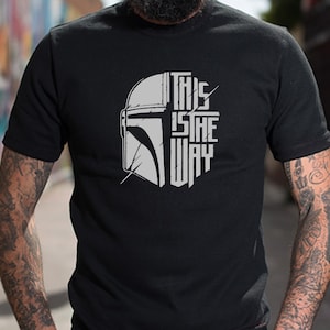Puede incluir: Camiseta negra con un gr&aacute;fico blanco de un casco mandaloriano y el texto "This is the way".