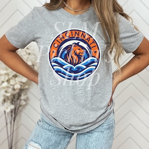 Puede incluir: Una camiseta gris con un gráfico circular que muestra la palabra "CINCINNATI" en naranja. El diseño incluye un león, olas azules y estrellas naranjas. La camiseta tiene cuello redondo y mangas cortas.