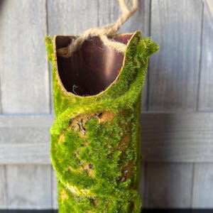 May include: A cylindrical decorative planter covered in vibrant green moss. The planter has a brown interior and a natural rope handle. The moss-covered exterior gives it a rustic, natural appearance, suitable for indoor or outdoor use.