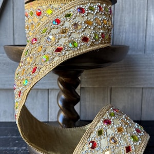 May include: A spool of gold ribbon with a decorative edge, embellished with colorful gemstones. The ribbon is displayed on a wooden stand, showcasing its intricate design and sparkling details. The ribbon is ideal for crafts and embellishments.