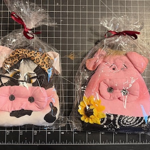 May include: Two stuffed fabric animal ornaments packaged in clear plastic bags with maroon ribbons. The left ornament is a black and white cow with a leopard print headband and glasses. The right ornament is a pink pig with a sunflower and bandana.