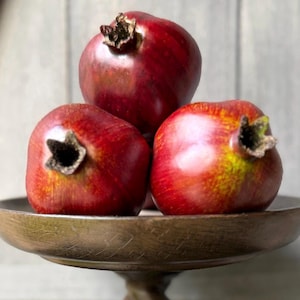 May include: Three whole pomegranates with deep red, smooth skins and prominent, dark brown calyxes at the top, arranged in a cluster on a dark brown, circular wooden pedestal stand with a smooth, polished finish and a sturdy, central base.