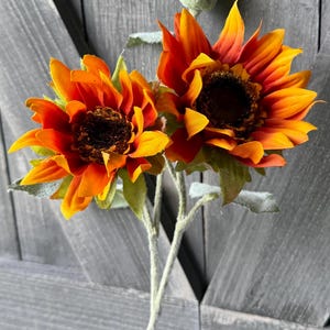 May include: Two artificial sunflowers with vibrant orange and yellow petals and dark brown centers. The stems are light green and the leaves are a muted green. A green bud is visible above the flowers. The background is a gray wooden surface.
