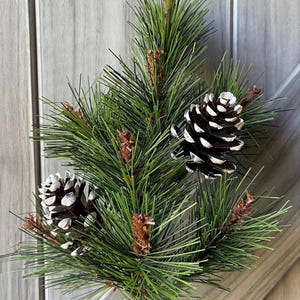 May include: A close-up of a faux pine branch with green needles and two pinecones. The pinecones are dark brown with white accents. The branch is a decorative item, perfect for holiday or seasonal decor.