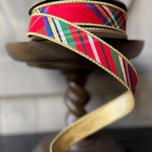 May include: A spool of fabric ribbon featuring a diagonal plaid pattern in red, green, white, blue, and black. The ribbon is edged with a metallic gold trim and has a solid gold-colored underside, creating a decorative and textured appearance.