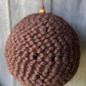 May include: A spherical ornament crafted from dark brown yarn, featuring a dense, textured crochet stitch pattern. A small, light-colored wooden bead is positioned at the top, where a thin brown cord extends upward for hanging purposes.