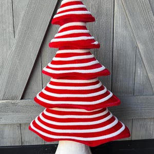 May include: A decorative tree-shaped ornament featuring horizontal red and white stripes. The structure consists of five stacked, tiered layers that taper toward the top, all supported by a solid white conical base at the bottom of the object.