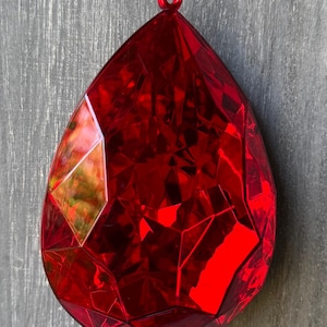 May include: A faceted, teardrop-shaped, red ornament with a gold hanging string. The ornament has a glossy finish and is designed with multiple flat surfaces that reflect light. The background is a gray wooden surface.