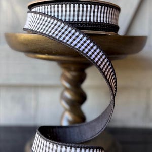 May include: A spool of fabric ribbon featuring a black and white gingham check pattern. The ribbon has solid black finished edges and is wound around a circular cardboard core, with a length of the ribbon cascading downward in a loose curve.