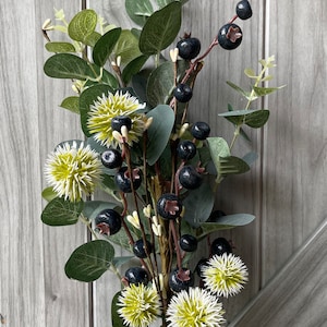 May include: A decorative floral arrangement featuring dark blue berries, green eucalyptus leaves, and spiky, light green and white flowers. The stems are brown and wrapped with a gold-colored material. The background is a light gray wooden surface.