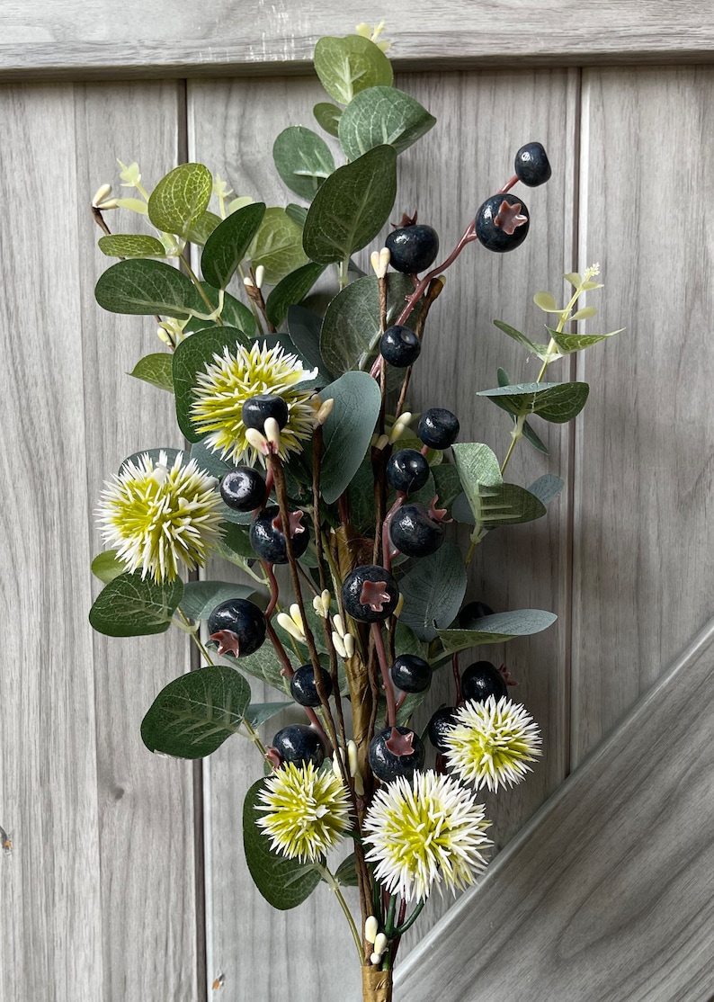May include: A decorative floral arrangement featuring dark blue berries, green eucalyptus leaves, and spiky, light green and white flowers. The stems are brown and wrapped with a gold-colored material. The background is a light gray wooden surface.
