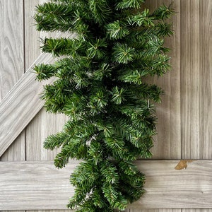 May include: A teardrop-shaped artificial evergreen swag featuring dense, realistic green pine needles. The foliage is arranged in a tapered, vertical design with multiple layered branches extending outward from a central spine, creating a full, lush appearance.