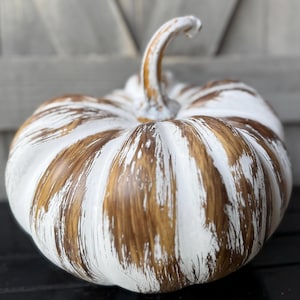 May include: A decorative pumpkin featuring a rounded, ribbed shape with a prominent stem. The surface is finished with a distressed, hand-painted effect, alternating between streaks of bright white and deep brown, creating a textured appearance.