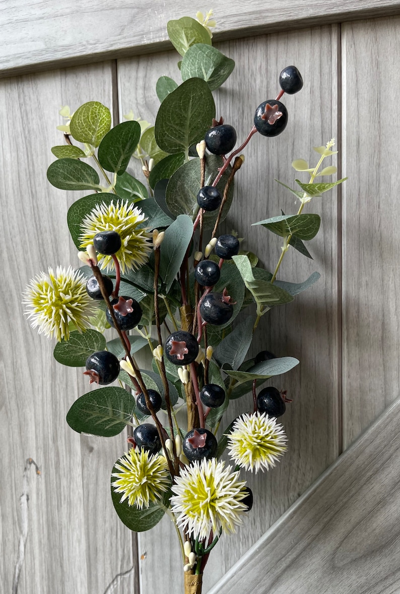 May include: Artificial floral arrangement featuring dark blue berries, green leaves, and spiky, light green and white flowers. The arrangement is set against a light gray wooden background, creating a natural aesthetic.
